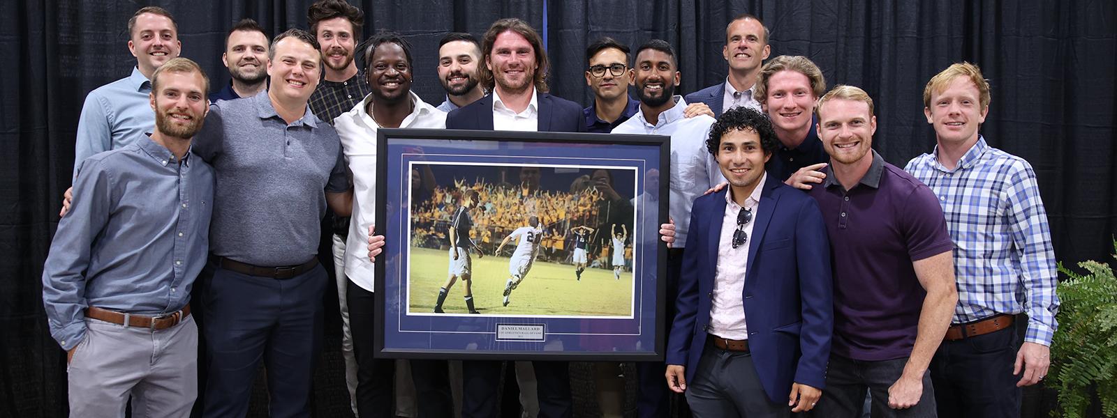 Daniel Mallard displays his Hall of Fame award with members of the 2012 men's soccer team. The photo shows Mallard celebrating after scoring the first goal in CIU history.)