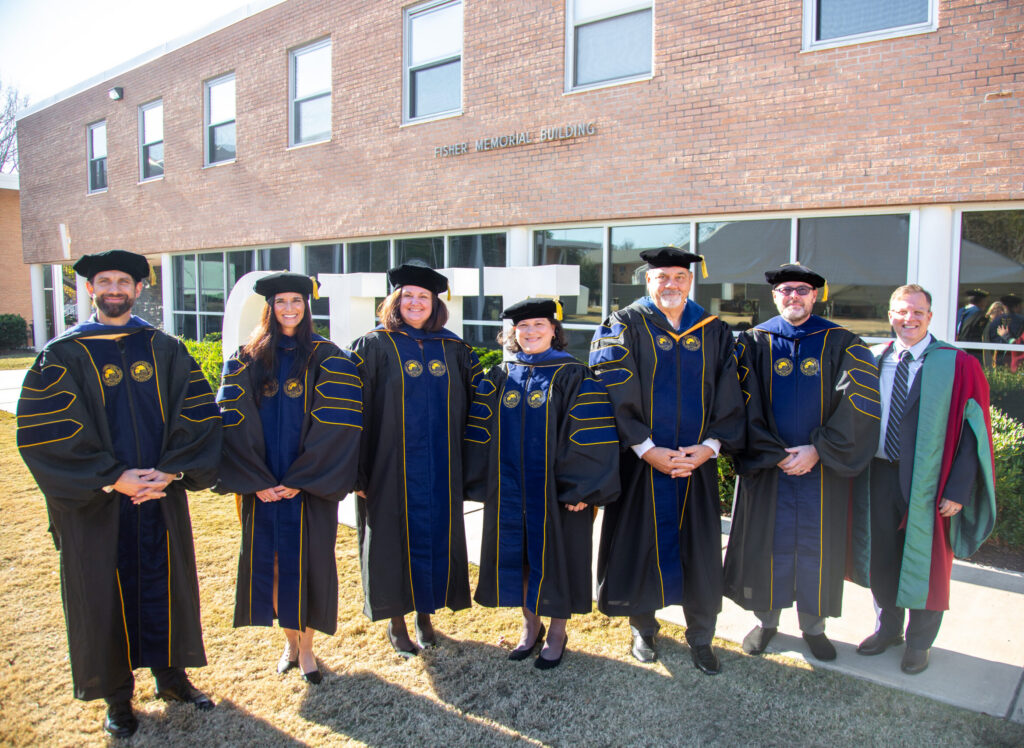 A group of doctoral graduates and faculty in academic regalia stand outside the Fisher Memorial Building on the CIU campus