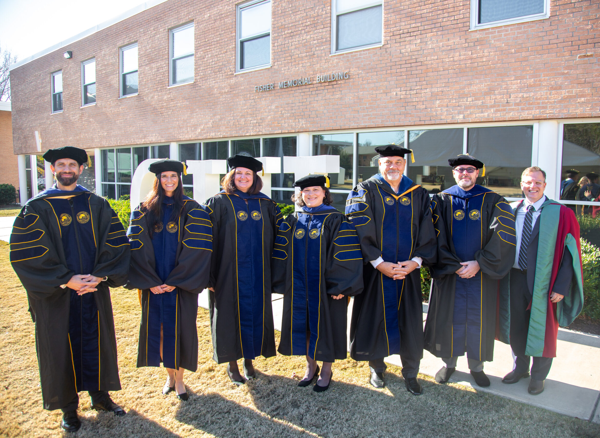 A group of doctoral graduates and faculty in academic regalia stand outside the Fisher Memorial Building on the CIU campus