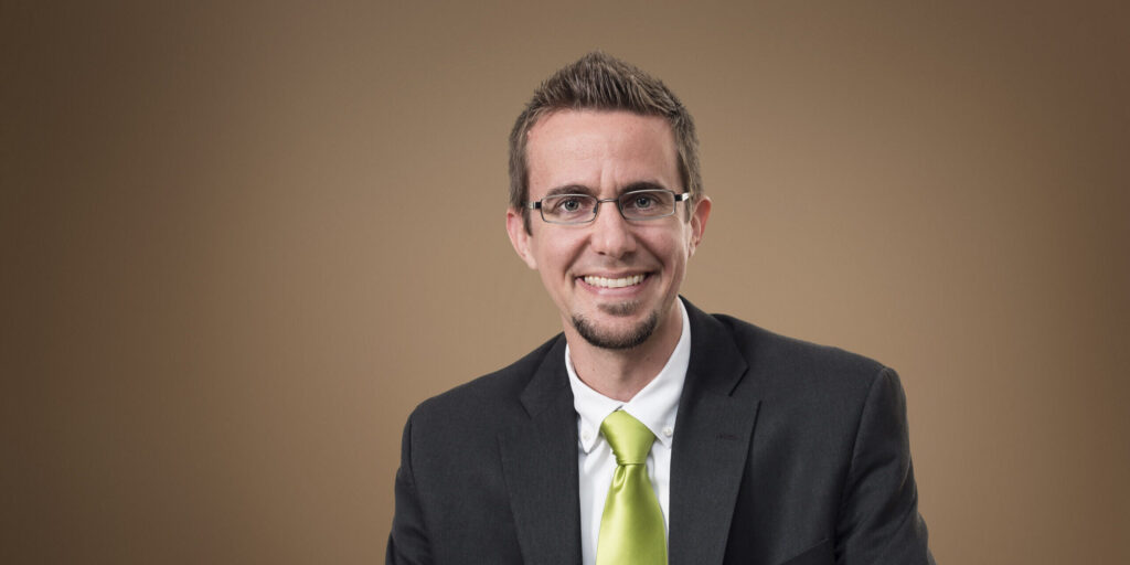 Dr. Nathaniel Jeanson, Columbia International University's first Visiting Research Professor, smiling in a dark suit and green tie against a neutral background.