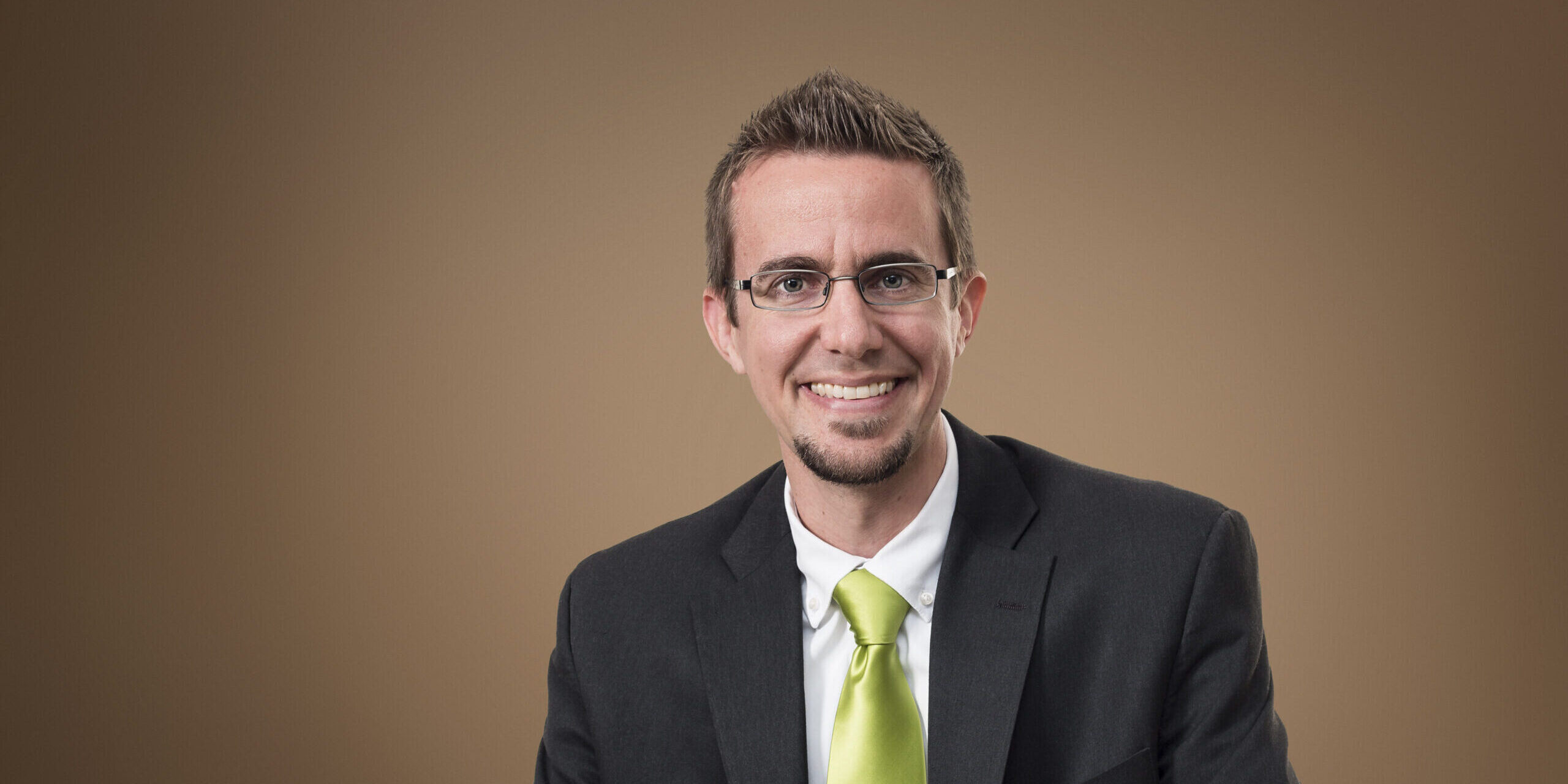 Dr. Nathaniel Jeanson, Columbia International University's first Visiting Research Professor, smiling in a dark suit and green tie against a neutral background.
